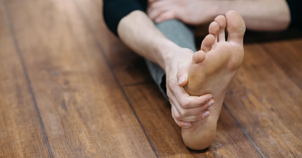 A man seated on a wooden floor with both hands wrapped around the arch of his bare foot.