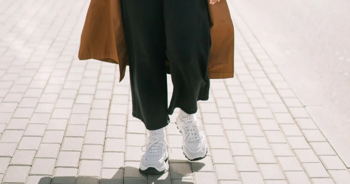 A pregnant woman walking on a paved sidewalk, seen from the knees down in white cushioned sneakers.