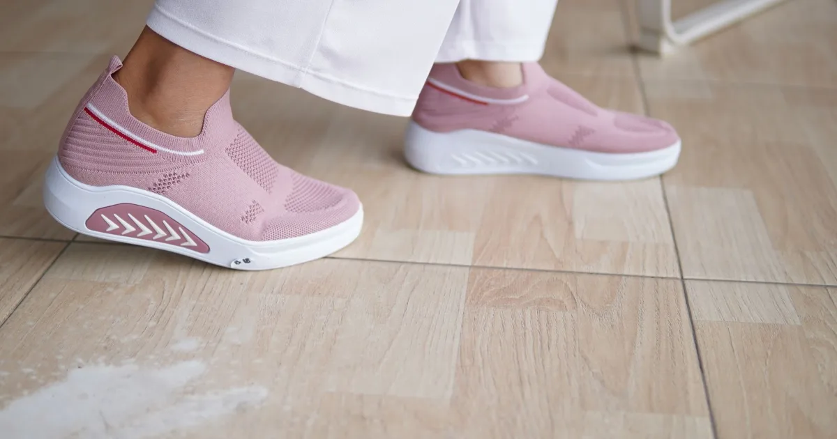 A pair of pink slip-on walking sneakers worn with cropped white trousers on a tiled floor.